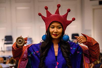 A young Asian person with long dark hair walks in a fashion show, wearing a bright pink, five-pointed felt hat, embroidered blue shirt and striped red jacket, oversize blue felt ball earrings, and an oversize gray circular ring. 