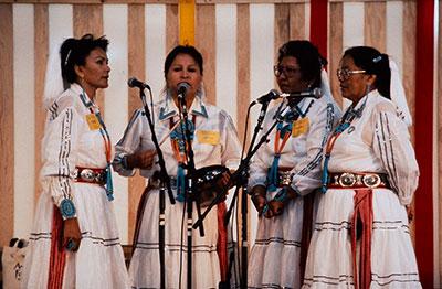 Four people in matching white dress and turquoise jewelry sing into microphones on stage.
