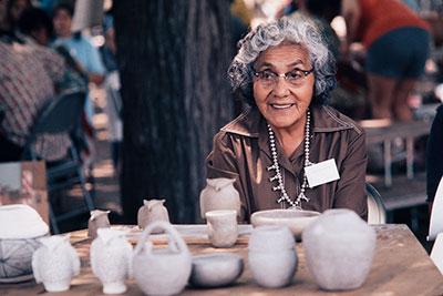 A woman smiles, sitting at a table with cream-colored ceramics, including small vases and owl figures.