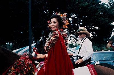 People ride on horseback, including a woman in red cape and flower lei and crown.