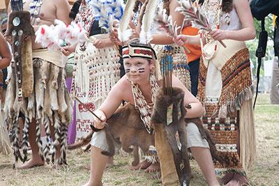 A young boy squats in an attack stance, holding wooden arrows, and wearing tribal dress.
