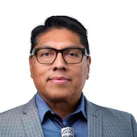On a pure white background, a man wearing a gray suit jacket, blue button-up shirt, and a silver bolo tie looks straight ahead.