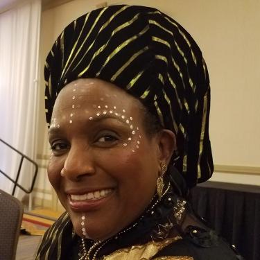 A woman smiles, wearing a black and gold striped head covering and white dotted face paint.