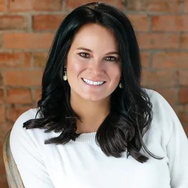 A smiling woman with long black hair wearing a white sweater sitting in front of a brick wall.