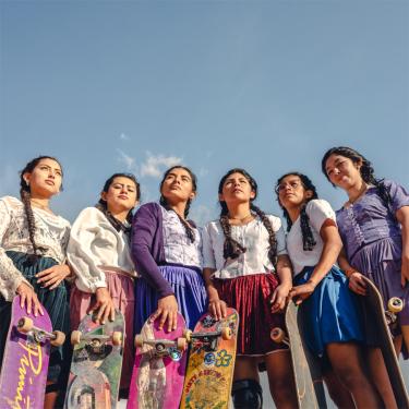Six women in colorful skirts stand holding their skateboards.