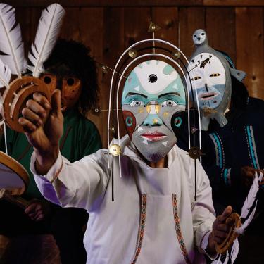 Three people in wooden humanoid masks holding drums and feather-adorned instruments.