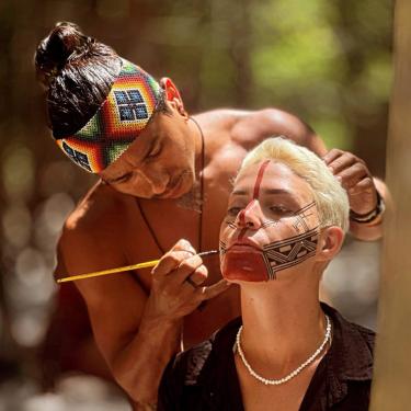 A man paints another person's face with red and black patterns.