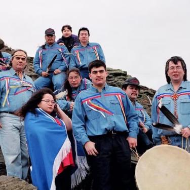 A group of people in matching denim tops adorned with blue and red ribbons across the chest pose on a rock face.