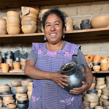 A woman holding a clay pot stands in front of shelves of pottery.