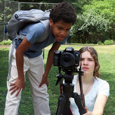 Two people crouch behind a camera on a tripod.