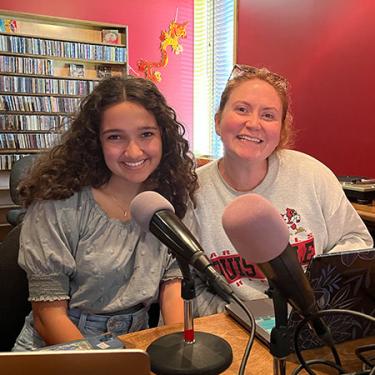 Two people smile sitting in front of microphones in a radio studio.