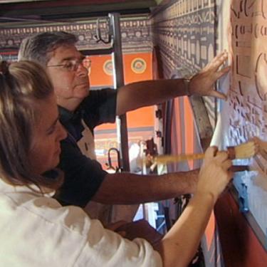 A man and a young woman use brushes on a stencil to embellish the moulding on a wall.