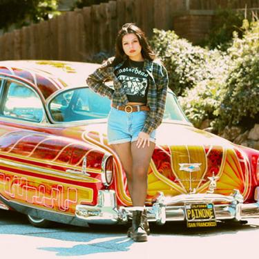 A woman in plaid overshirt and jean shorts, one hand on her hip, poses in front of an intricately painted red and yellow lowrider car.