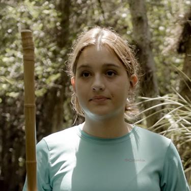 A young person hikes through the woods with a long walking stick.