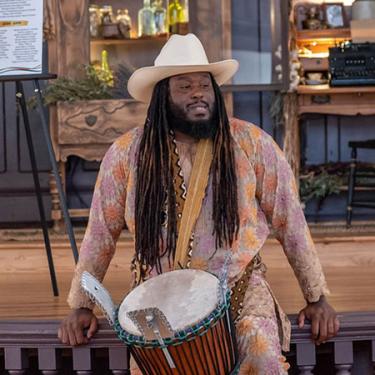 A man with long dark locs and beard, wearing a tan cowboy hat and pale purple and orange outfit, sits at the front of a stage with a hand drum slung over his shoulder.