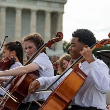 Teens in white dress shirts play cellos outdoors with the Lincoln Memorial in the background.