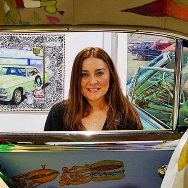 Through the open windows of a car with flying sandwiches painted in the interior, a woman with long brown hair smiles.