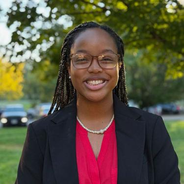 A young woman with long dark braids, glasses, and black blazer over a fucshia blouse smiles.