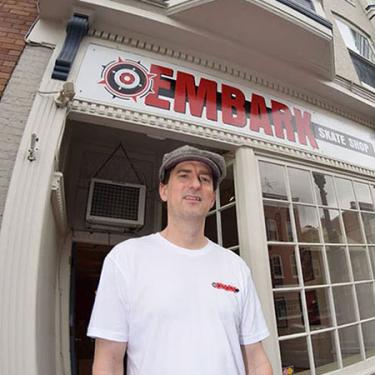 A man poses outside a shope with sign above the door: Embark Skate Shop.