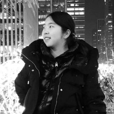 A young woman with dark hair and heavy black jacket smiles, a nighttime cityscape behind her.
