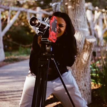 A young person crouches behind a camera on a tripod, squinting to see through the viewfinder.