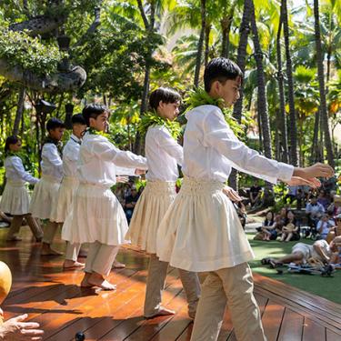 Boys in white perform hula on an outdoor stage in front of a crowd and palm trees.