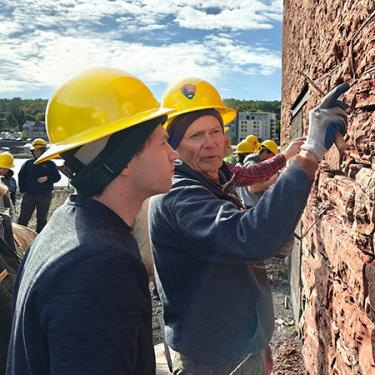 A man points to a detail in a red stone wall as a young person observes. Both wear yellow hard hats.
