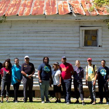 A group of people pose in a line in front of an old building with rusted red roof.