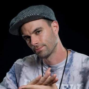 A man with a pageboy cap and tie-dye shirt poses.