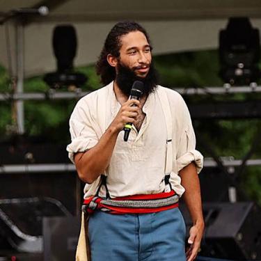 A man with dark hair in a ponytail and bushy beard speaks into a microphone in front of an outdoor stage.