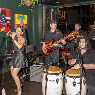 A band all dressed in black performs in a club, including a singer, bassist, and conga player.
