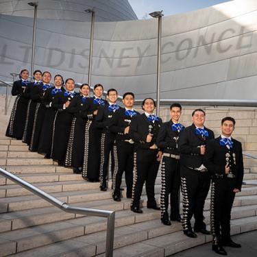 A group in matching black suits with blue ties pose descending a staircase in from of Disney Concert Hall.