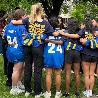 Teenage students huddle in a circle outdoors wearing various blue jerseys. Two of their shirts read Class of 2025.