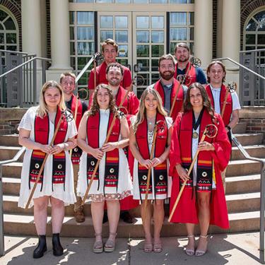 Ten young adults pose in front of a brick building in red graduation stoles, each holding a lacrosse stick.