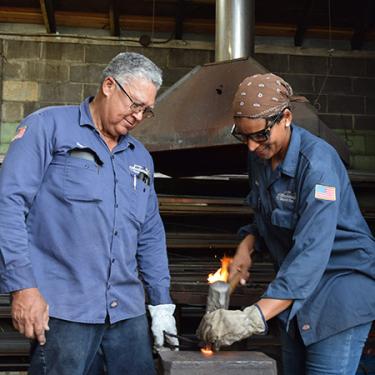 Two blacksmiths in navy work shirts form a piece of red-hot metal. One holds it in place while the other shapes it with a mallet.