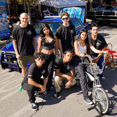 Seven teens pose next to rows of lowrider bicycles and cars parked in a street.