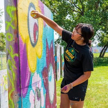 A young woman spraypaints a makeshift wooden wall outside, using bright yellow, purples, and turquoise.