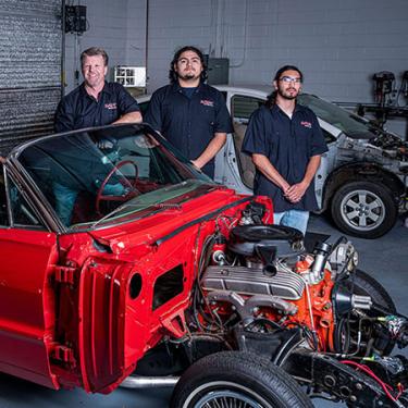 People in matching black work shirts pose around a bright red convertible car with the hood removed to show the engine.
