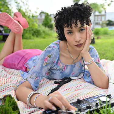 A woman lays on a blanket in the grass, holding her head propped up with one hand and fiddling with a drum machine with the other.