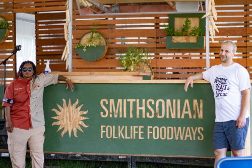 A woman in a half beige, half red chef's coat and man in a white Words, Beats & Life Inc. t-shirt pose and smile with a green Smithsonian Folklife Foodways sign.