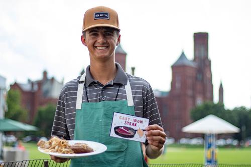 A teenage boy poses and smiles while holding a plate of cast-iron steak in one hand and an illustrated recipe card in the other; the Smithsonian Castle is in the background.