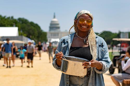 A woman wearing yellow-tinted sunglasses and posing on the National Mall with a metal pot in her hands; the Capitol Building is in the background.