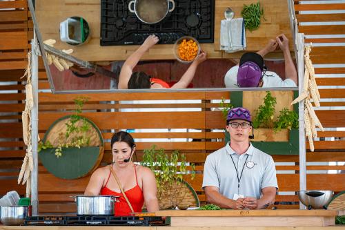 A woman and man demonstratea recipe on Foodways' stage; the woman prepares to put ingredients into a metal pot with boiling soup broth; the man speaks to the audience.