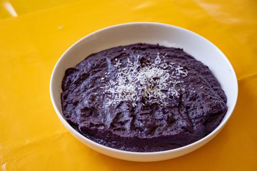 A bowl of deep-purple sweet potato pudding sprinkled in coconut shreds rests on a bright yellow table.