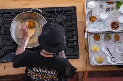 A man wearing a black bucket hat and black graphic tee mixes seasonings in  a silver pot using a spoon and wooden spatula; a colleciton of seasonings and other ingredients are displayed next to the stove.