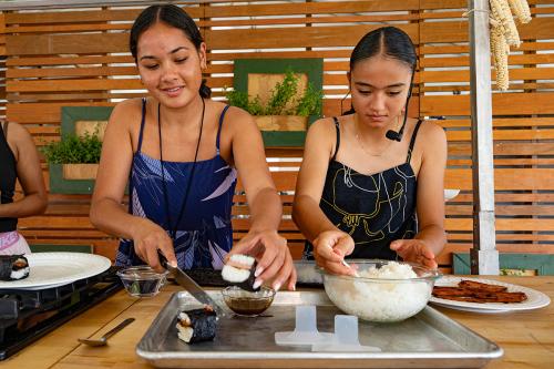 Two young girls work together to make spam musubi; one girl cuts a roll of spam musubi in half; the other girl scoops a spoonful of white rice from a glass bowl.