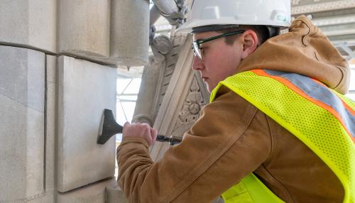 A young woman wearing a hard hat, safety glasses, yellow safety vest, and tan jacket uses a metal tool on a white stone of a building.