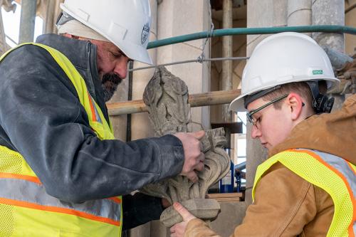 Two people in hard hats and safety vests move a carved stone ornament.