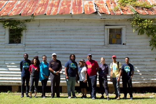 A group of people pose in a line in front of an old building with rusted red roof.