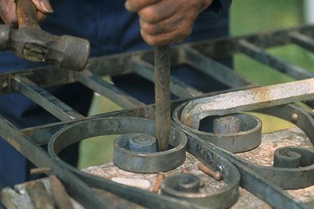 Close-up on a blacksmith shaping a curling ornamentation in a metal gate using two metal tools.
